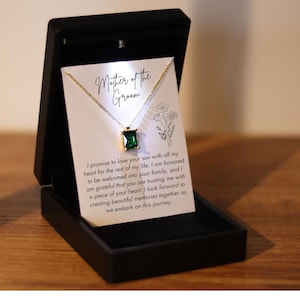 May include: A white card with the text "Mother of the Groom" and a message about love and family. A gold necklace with a green gemstone pendant is displayed on the card.
