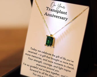 May include: A gold-colored necklace with a square, emerald-green gemstone pendant. The necklace is displayed on a card that reads "On Your Transplant Anniversary" with a heartfelt message. The necklace is presented in a black box.