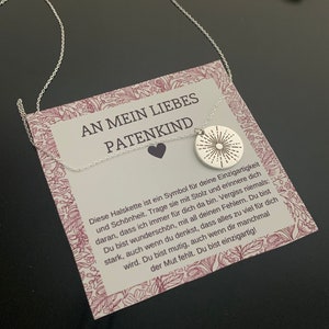 May include: A silver necklace with a small silver pendant featuring a sunburst design. The necklace is displayed on a card with the text "An mein liebes Patenkind" and a message about the necklace being a symbol of uniqueness and beauty.