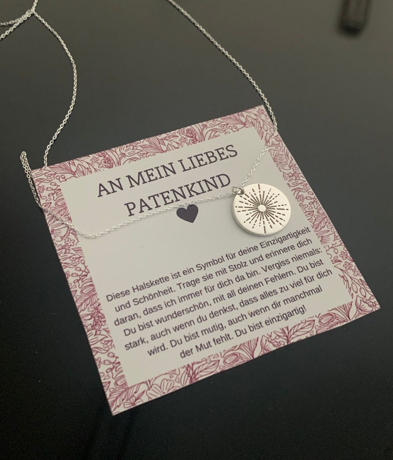 May include: A silver necklace with a small silver pendant featuring a sunburst design. The necklace is displayed on a card with the text "An mein liebes Patenkind" and a message about the necklace being a symbol of uniqueness and beauty.