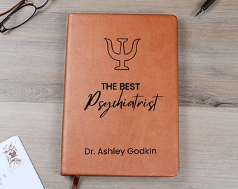 May include: A brown leather journal with a black engraved symbol and the text "The Best Psychiatrist Dr. Ashley Godkin".