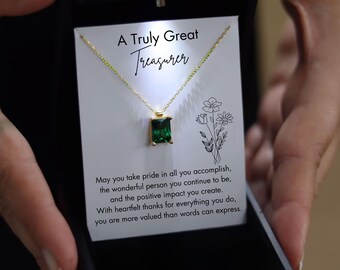 May include: A gold-colored necklace with a rectangular, emerald-green pendant is displayed in a black gift box. The necklace is presented on a card with the text "A Truly Great Treasurer" and a heartfelt message. The box is held by a person.