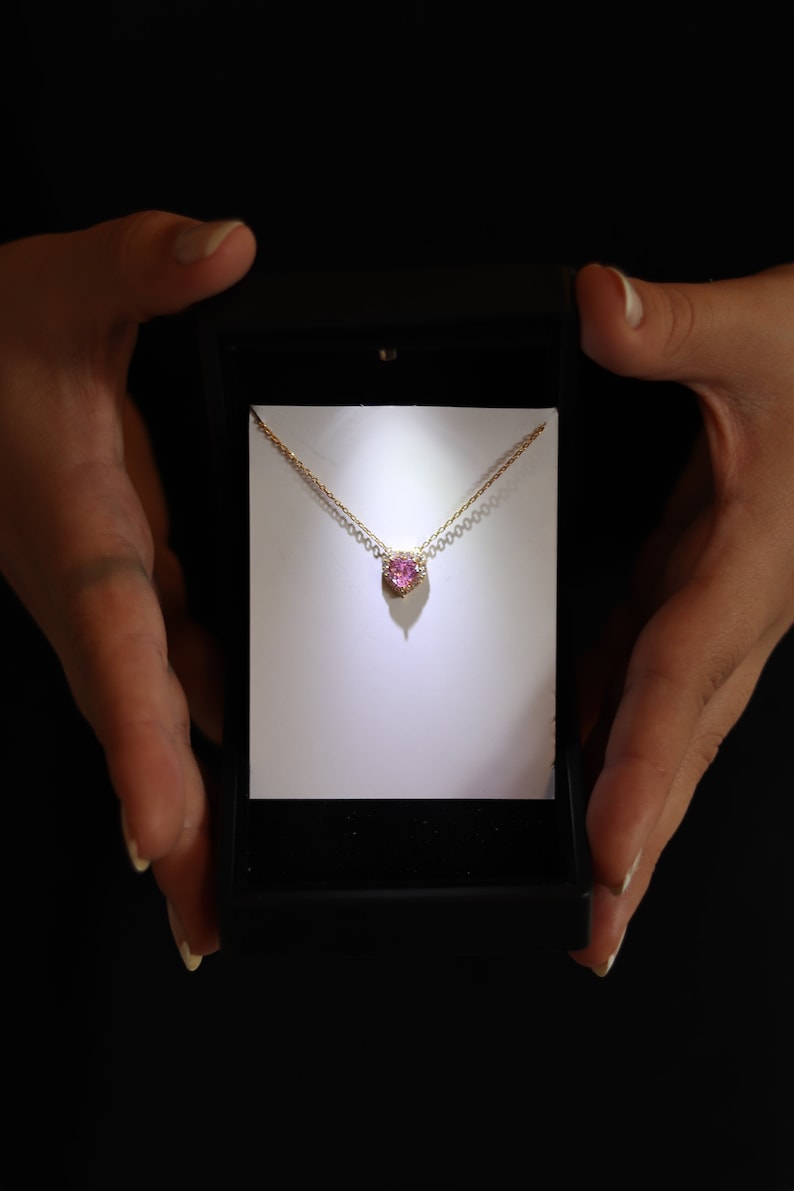 May include: A gold chain necklace with a pink heart-shaped gemstone pendant. The pendant is surrounded by smaller diamonds.