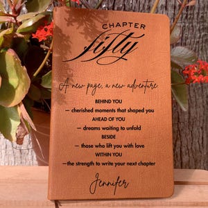 May include: A brown leather journal with the words "Chapter Fifty" and "A new page, a new adventure" printed on the cover. The journal includes inspirational text and the name "Jennifer" at the bottom. The journal is displayed in front of a plant.