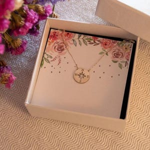 May include: A gold-colored compass pendant necklace displayed in an open white gift box. The box contains a card with floral designs. A bouquet of pink and purple flowers is in the background. The necklace is on a white background.