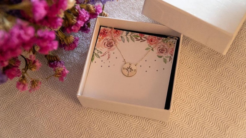 May include: A gold-colored compass pendant necklace displayed in an open white gift box. The box contains a card with floral designs. A bouquet of pink and purple flowers is in the background. The necklace is on a white background.