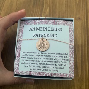 May include: A small gift box with a gold necklace inside. The necklace has a round pendant with a sunburst design. The box has a white background with pink floral accents and the text "An mein liebes Patenkind" in black lettering. The text reads: "Diese Halskette ist ein Symbol f&uuml;r deine Einzigartigkeit und Sch&ouml;nheit. Trage sie mit Stolz und erinnere dich daran, dass ich immer f&uuml;r dich da bin. Vergiss niemals: Du bist wundersch&ouml;n, mit all deinen Fehlern. Du bist stark, auch wenn du denkst, dass alles zu viel f&uuml;r dich wird. Du bist mutig, auch wenn dir manchmal der Mut fehlt. Du bist einzigartig!"