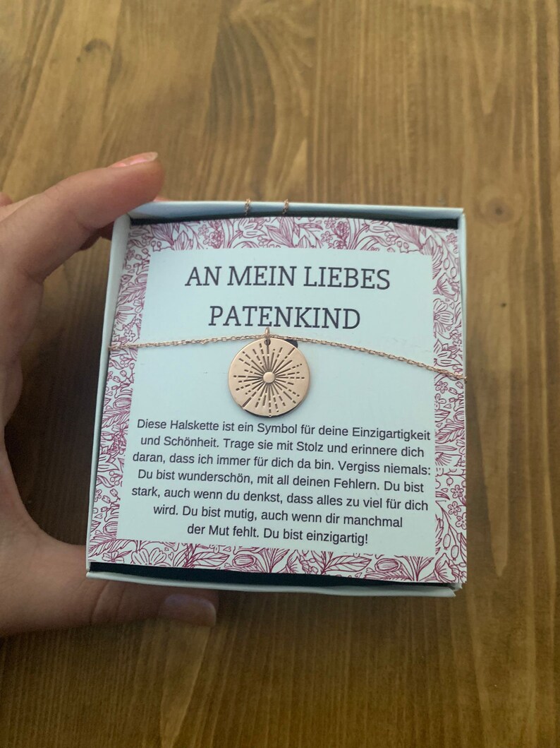 May include: A small gift box with a gold necklace inside. The necklace has a round pendant with a sunburst design. The box has a white background with pink floral accents and the text "An mein liebes Patenkind" in black lettering. The text reads: "Diese Halskette ist ein Symbol f&uuml;r deine Einzigartigkeit und Sch&ouml;nheit. Trage sie mit Stolz und erinnere dich daran, dass ich immer f&uuml;r dich da bin. Vergiss niemals: Du bist wundersch&ouml;n, mit all deinen Fehlern. Du bist stark, auch wenn du denkst, dass alles zu viel f&uuml;r dich wird. Du bist mutig, auch wenn dir manchmal der Mut fehlt. Du bist einzigartig!"