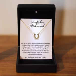 May include: A gold-colored horseshoe pendant necklace displayed in a black gift box. The necklace is on a white card with the German text "Herzlichen Gl&uuml;ckwunsch" and a message of congratulations.