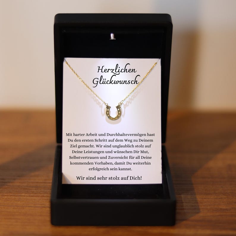 May include: A gold-colored horseshoe pendant necklace displayed in a black gift box. The necklace is on a white card with the German text "Herzlichen Gl&uuml;ckwunsch" and a message of congratulations.