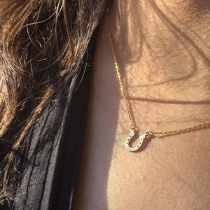 May include: A gold-colored horseshoe pendant necklace. The horseshoe charm has small, dark accents and the word "Cursive" engraved on it. The necklace is worn around the neck, with the chain visible.