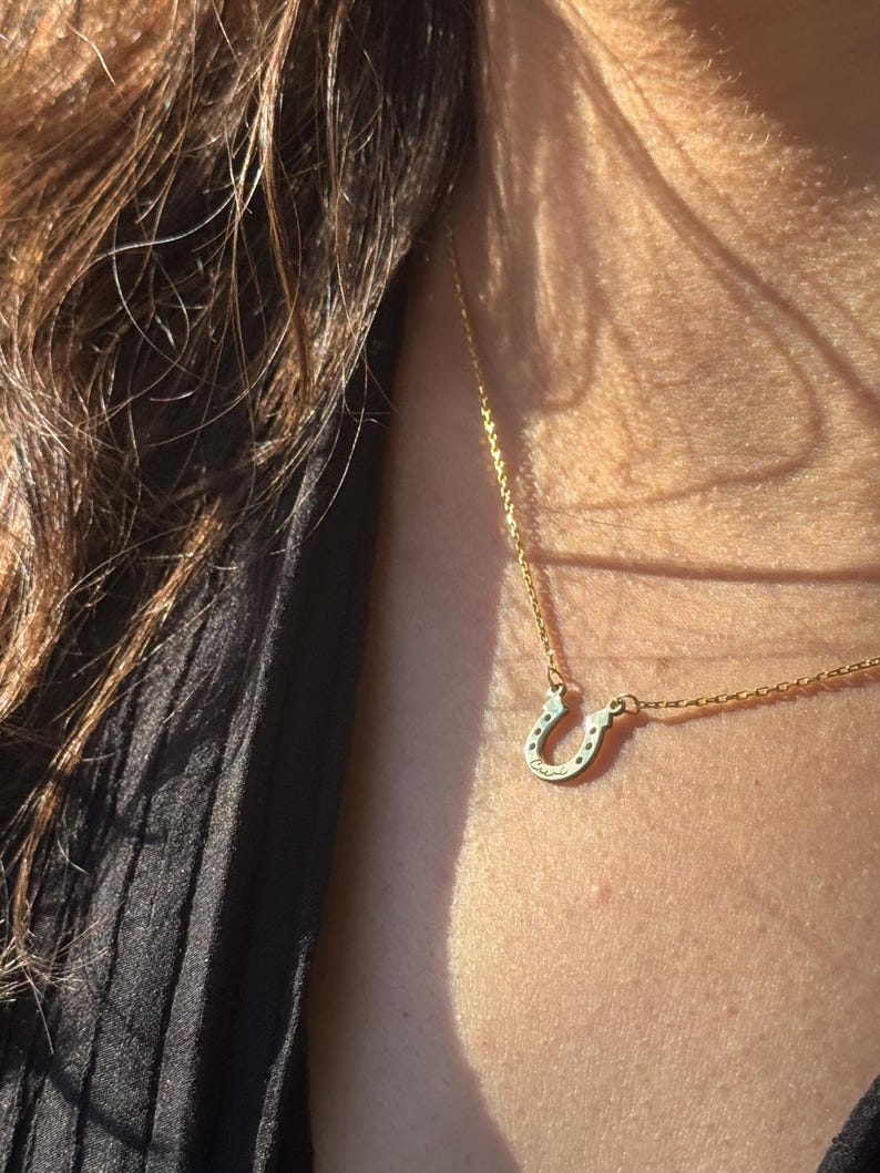 May include: A gold-colored horseshoe pendant necklace. The horseshoe charm has small, dark accents and the word "Cursive" engraved on it. The necklace is worn around the neck, with the chain visible.