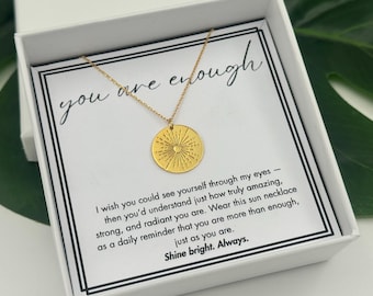 May include: A gold-colored sunburst pendant necklace on a delicate chain, presented in a white gift box. The card inside reads "you are enough" and includes an inspirational message. The necklace is a thoughtful gift.