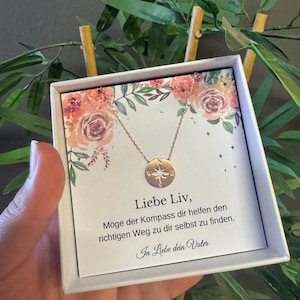 Peut inclure: Un collier pendentif boussole en or rose présenté dans un coffret blanc avec des motifs floraux. Le coffret contient du texte en allemand. Le collier a un pendentif circulaire avec des détails de boussole.
