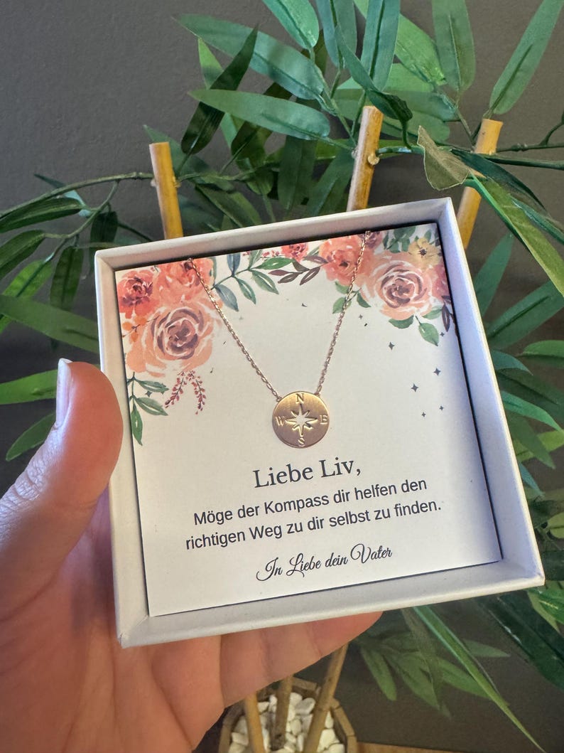 May include: A rose gold compass pendant necklace displayed in a white gift box with floral accents. The box contains text in German. The necklace features a circular pendant with compass details.