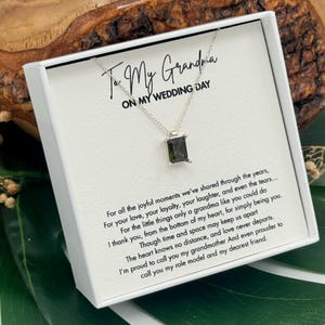 Può includere: Una collana d'argento con un pendente rettangolare verde scuro, presentata in un cofanetto bianco. La scatola riporta la scritta "To My Grandma ON MY WEDDING DAY" e un messaggio sentito. Lo sfondo comprende una superficie in legno e foglie verdi.