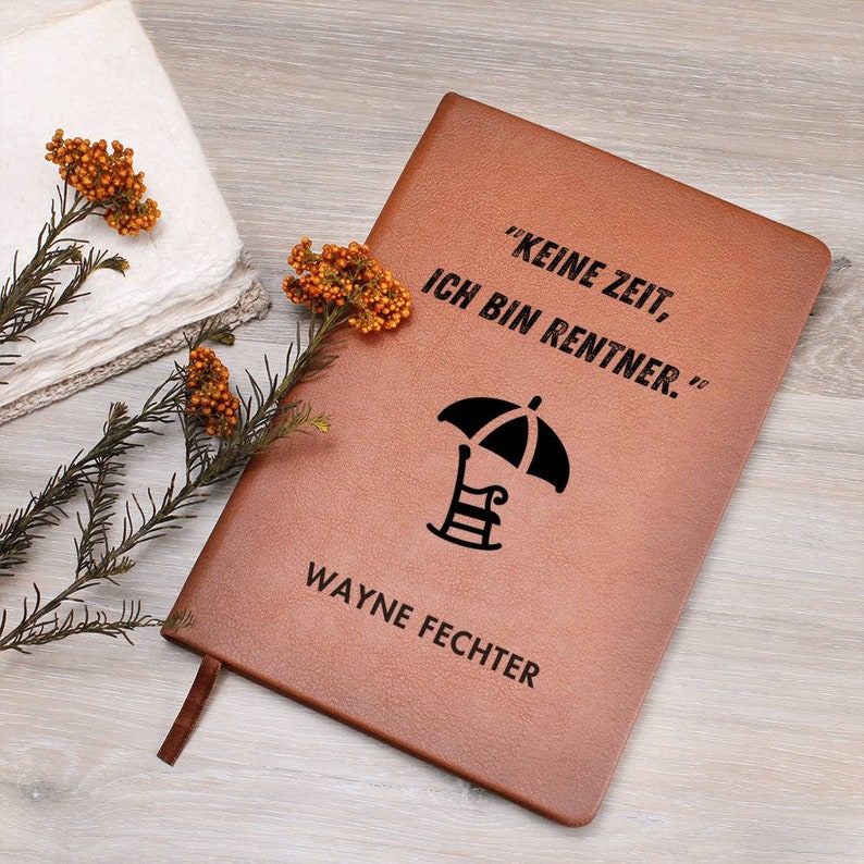 May include: Brown leather journal with the text "Keine Zeit, Ich Bin Rentner." and a black silhouette of a beach chair under an umbrella. The name "WAYNE FECHTER" is printed below the image.