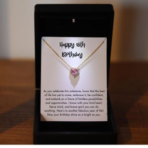 May include: A gold-colored necklace with a pink heart-shaped pendant, presented in a black gift box. The card inside reads "Happy 18th Birthday" and includes a message. A thoughtful gift for a special occasion.