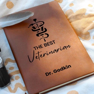 May include: Brown leather journal with a black engraved image of a snake wrapped around a staff with a cross at the top. The text "THE BEST VETERINARIAN Dr. Godkin" is engraved below the image.