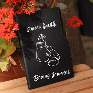 Może przedstawiać: Czarny skórzany dziennik z tekstem "Boxing Journal" i białą ilustracją bokserskich rękawic. Imię "James Smith" jest napisane na okładce białym kolorem.