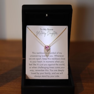May include: A gold necklace with a pink heart pendant set in a clear crystal halo. The necklace is in a black box with a white card that reads "To My Brave Military Daughter." This necklace is a symbol of my unwavering love for you. Whenever we are apart, keep this necklace close to your heart. In moments when you feel like it's just you against the world, or when challenging times come your way, remember this: You are deeply loved by your family, and we will always stand by your side.