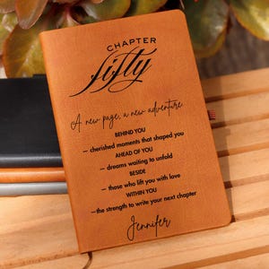 以下が含まれることがあります： 「Chapter Fifty」と「A new page, a new adventure」の文字がプリントされた、茶色の革製ジャーナル。インスピレーションを与えるテキストと、下部に「Jennifer」という名前が記載されています。