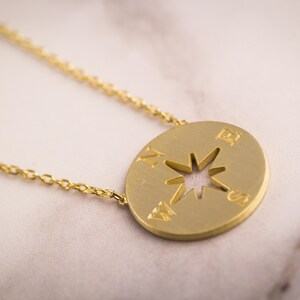 May include: A gold-colored necklace featuring a compass pendant. The pendant has a cut-out star design in the center and directional markings. The necklace chain is delicate and gold, resting on a marble surface.