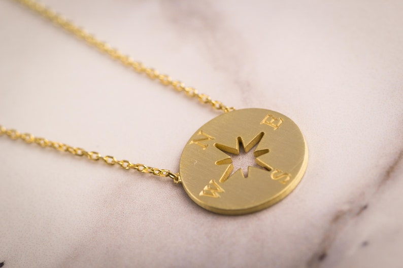 May include: A gold-colored necklace featuring a compass pendant. The pendant has a cut-out star design in the center and directional markings. The necklace chain is delicate and gold, resting on a marble surface.