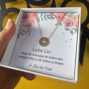 May include: A rose gold compass pendant necklace in a white gift box. The necklace features a circular pendant with a compass design. The box has a floral design and German text.