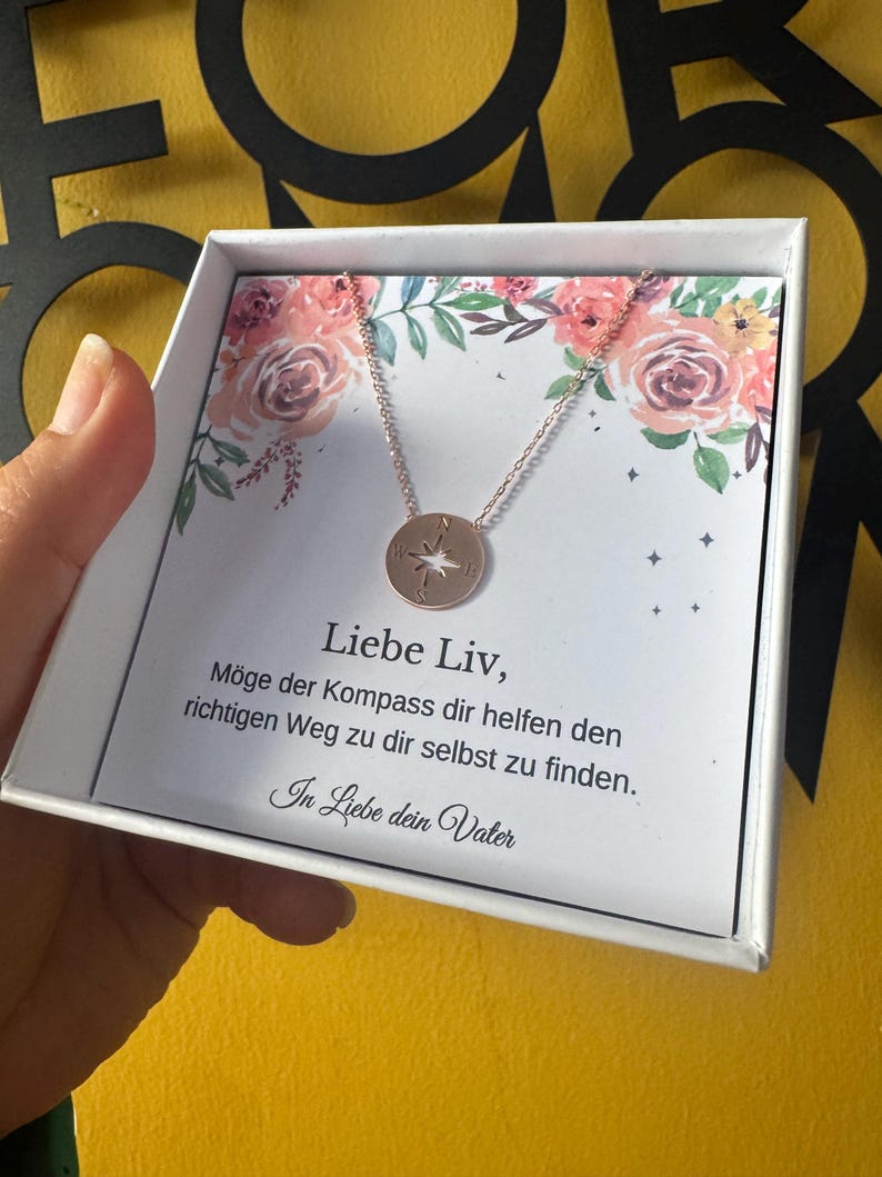 May include: A rose gold compass pendant necklace in a white gift box. The necklace features a circular pendant with a compass design. The box has a floral design and German text.