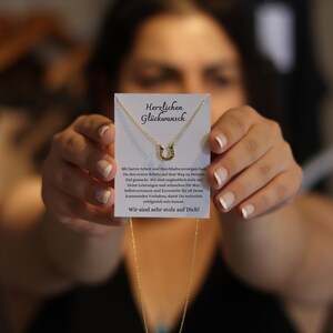 May include: A gold-colored horseshoe pendant necklace is displayed on a white card with German text. The card is held up by a person. The text on the card reads "Herzlichen Gl&uuml;ckwunsch" and a message of congratulations.
