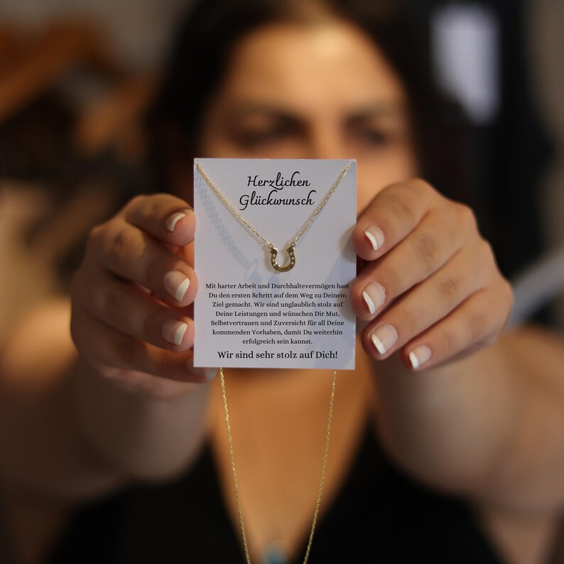 May include: A gold-colored horseshoe pendant necklace is displayed on a white card with German text. The card is held up by a person. The text on the card reads "Herzlichen Gl&uuml;ckwunsch" and a message of congratulations.