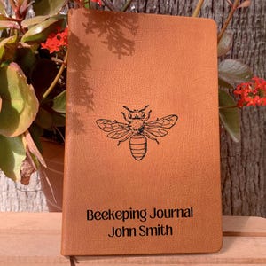 Könnte beinhalten: Ein braunes, ledergebundenes Tagebuch mit einer Bienenillustration und dem Text "Beekeeping Journal John Smith" auf dem Einband.