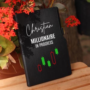 Könnte beinhalten: Schwarzes Ledernotizbuch mit dem Text "Christian Millionaire in Progress" und einem grün-roten Candlestick-Chart.