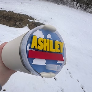Peut inclure: Un gobelet de voyage blanc avec un couvercle bleu, rouge et jaune avec le nom "ASHLEY" en lettres jaunes. Le couvercle a un motif de nuage.