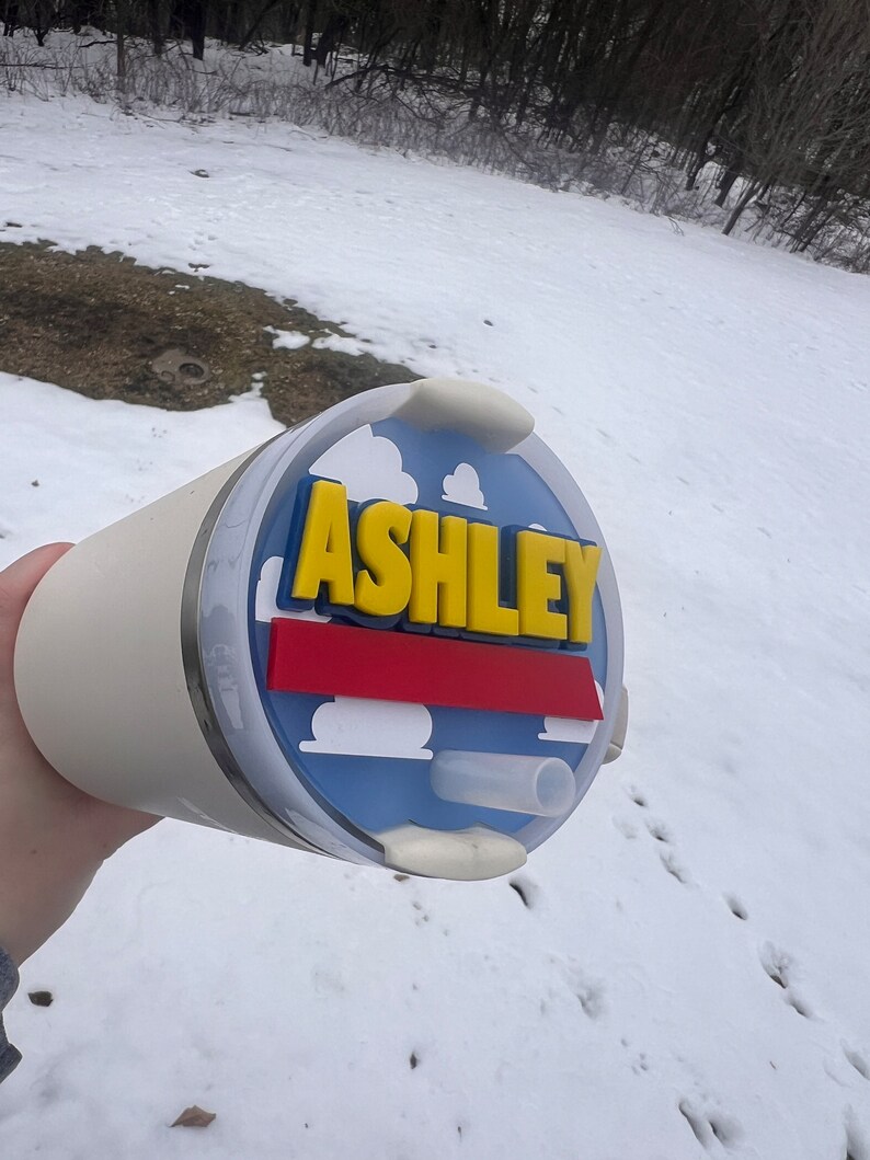 Peut inclure: Un gobelet de voyage blanc avec un couvercle bleu, rouge et jaune avec le nom "ASHLEY" en lettres jaunes. Le couvercle a un motif de nuage.