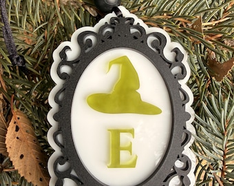 Puede incluir: Un adorno decorativo enmarcado en blanco y negro con un sombrero de bruja y la letra "E" en verde. El adorno cuelga de un cordón negro y se encuentra sobre un fondo de agujas de pino verdes.