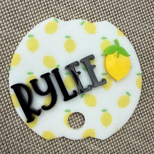 May include: A white car coaster featuring a lemon pattern and the name "RYLEE" in black lettering. A yellow lemon with a green leaf is on the right. The coaster has a scalloped edge and a small hole.