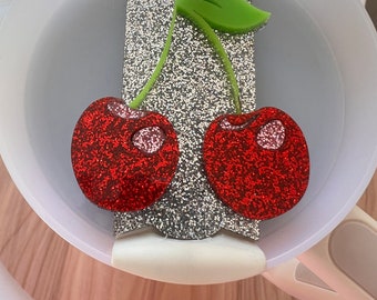 May include: A pair of red glitter cherry earrings with green stems and leaves on a silver glitter background.