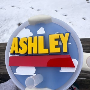 Peut inclure: Un couvercle de tasse personnalis&eacute; bleu et blanc avec une bande rouge et des nuages blancs. Le nom "ASHLEY" est &eacute;crit en lettres 3D jaunes sur le couvercle.