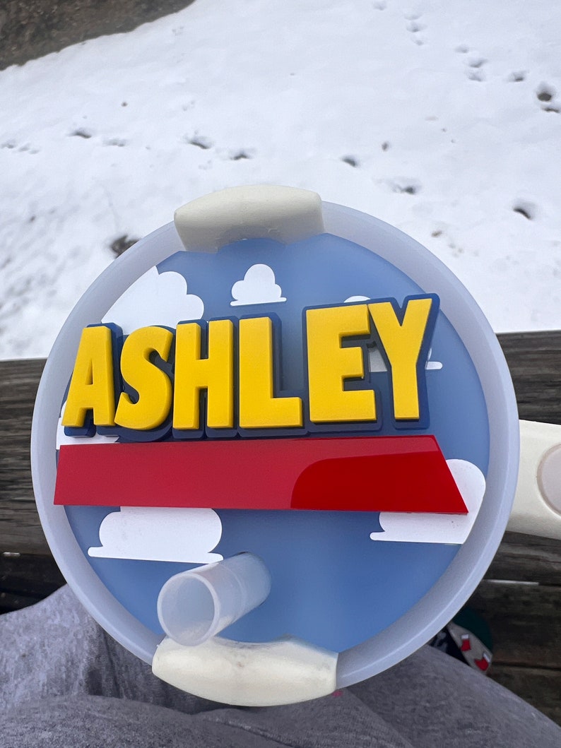 Peut inclure: Un couvercle de tasse personnalis&eacute; bleu et blanc avec une bande rouge et des nuages blancs. Le nom "ASHLEY" est &eacute;crit en lettres 3D jaunes sur le couvercle.