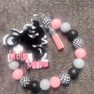 May include: A black and white checkered keychain with a pink tassel and a black silhouette of a dirt bike rider with the text "Moto Mama". The keychain is attached to a beaded bracelet with black, white, pink, and gray beads.