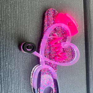 Pu&ograve; includere: Porta-stetoscopio in glitter rosa con un cuore e un design a stetoscopio. Il cuore ha la scritta "Nursy" all'interno.