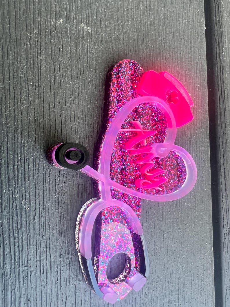 Pu&ograve; includere: Porta-stetoscopio in glitter rosa con un cuore e un design a stetoscopio. Il cuore ha la scritta "Nursy" all'interno.
