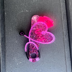 Pu&ograve; includere: Cuore rosa glitter con la parola "Nurse" all'interno, un cappello da infermiera rosa in cima e uno stetoscopio nero attaccato al lato.