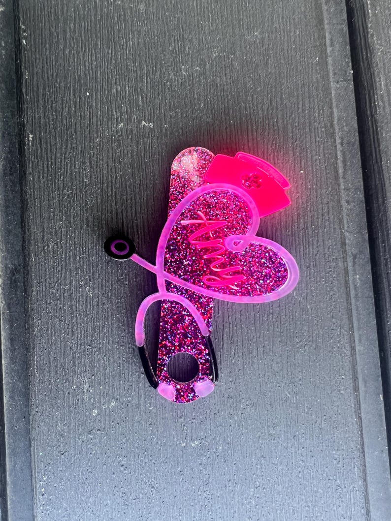 Pu&ograve; includere: Cuore rosa glitter con la parola "Nurse" all'interno, un cappello da infermiera rosa in cima e uno stetoscopio nero attaccato al lato.