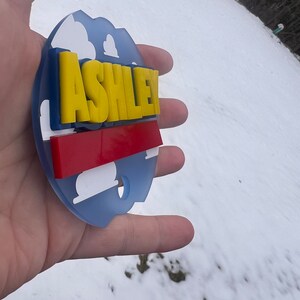 Peut inclure: Une d&eacute;coration ronde bleue et blanche avec une bande rouge et blanche et le nom "ASHLEY" en lettres jaunes.