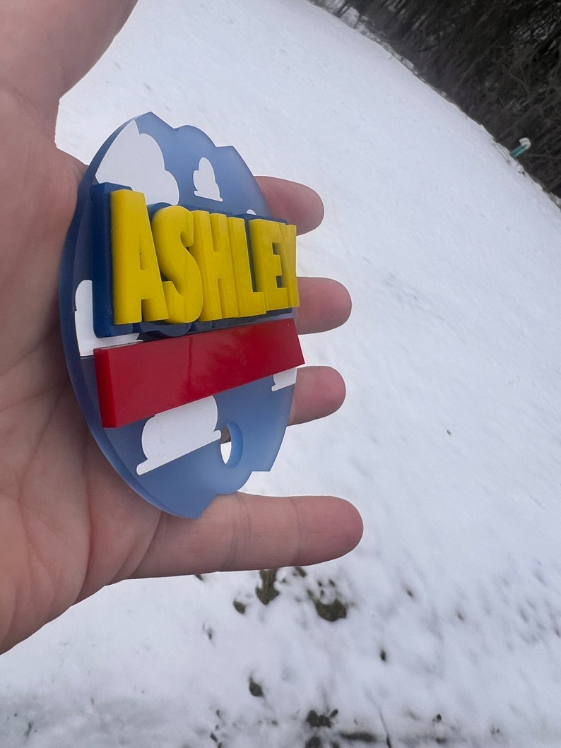 Peut inclure: Une d&eacute;coration ronde bleue et blanche avec une bande rouge et blanche et le nom "ASHLEY" en lettres jaunes.