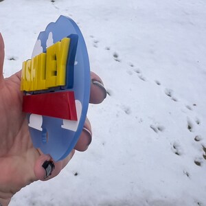 Peut inclure: Une &eacute;tiquette de nom personnalis&eacute;e bleue, jaune et rouge avec le nom "ASHLEY" en lettres jaunes avec un contour bleu. L'&eacute;tiquette de nom a la forme d'un nuage avec un fond blanc.