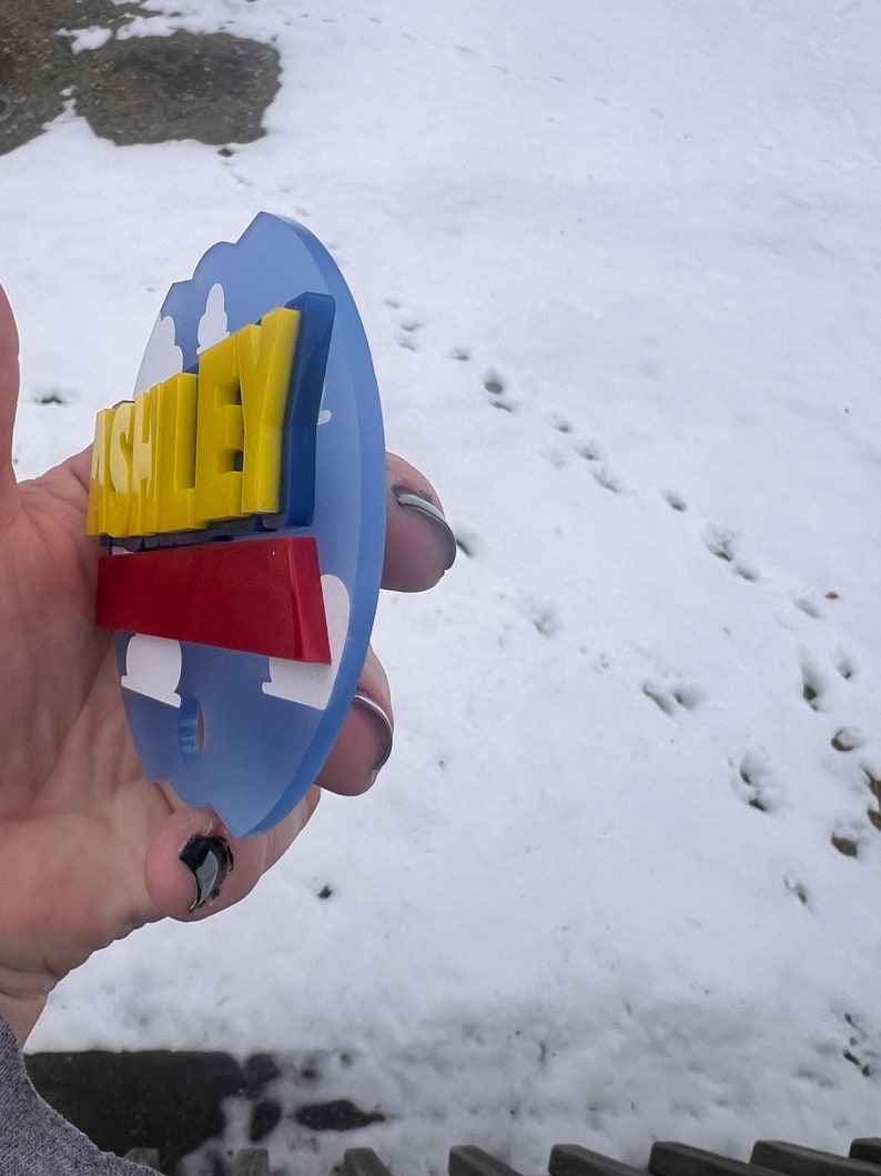 Peut inclure: Une &eacute;tiquette de nom personnalis&eacute;e bleue, jaune et rouge avec le nom "ASHLEY" en lettres jaunes avec un contour bleu. L'&eacute;tiquette de nom a la forme d'un nuage avec un fond blanc.